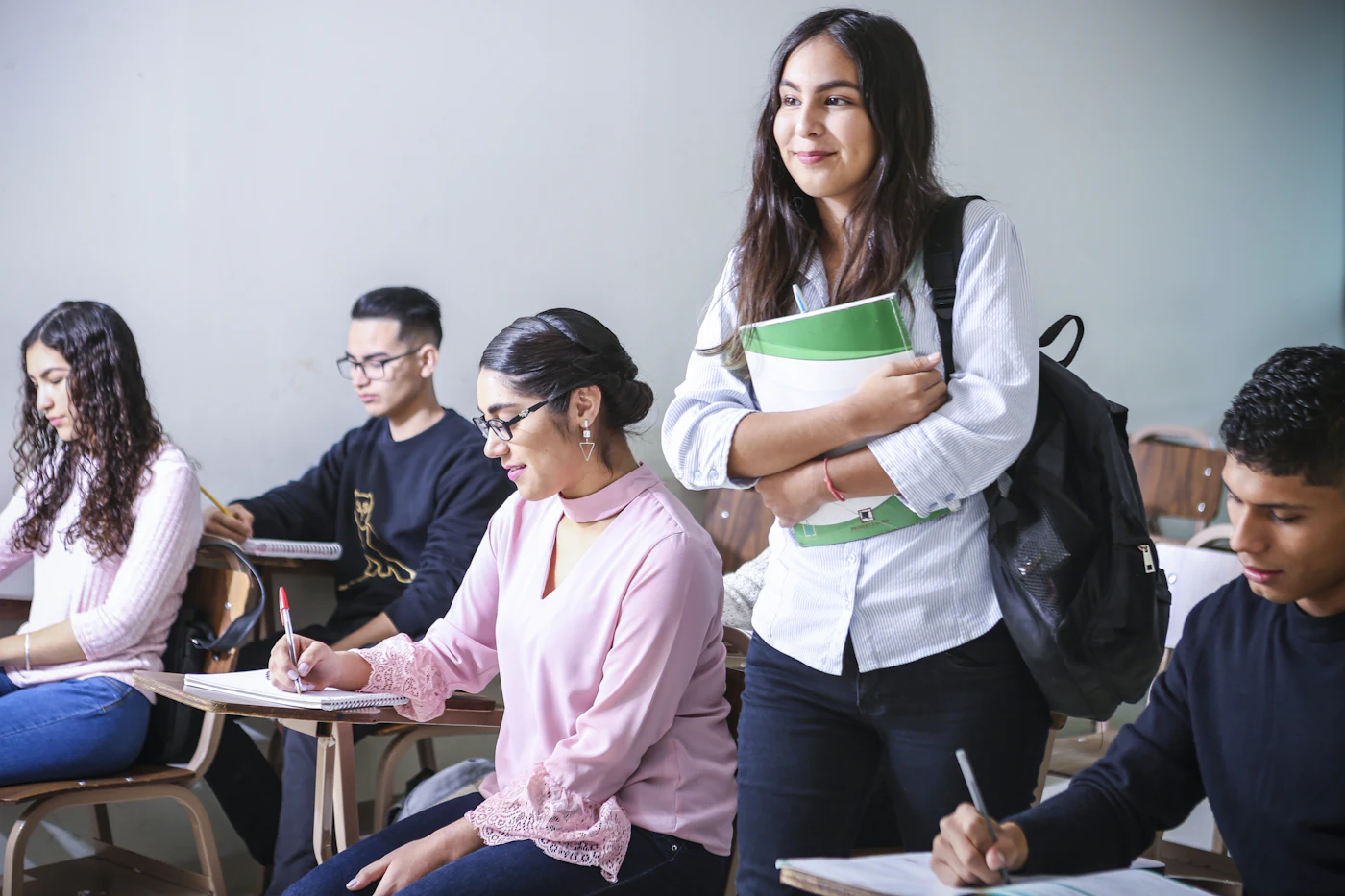 College students in a classroom; one stands smiling with a textbook and backpack